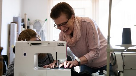 An older woman teaches a younger one how to sew on a machine. She smiles. 