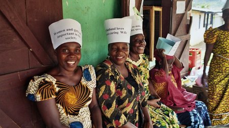 Three women smiling at the camera and wearing feminist messages on headwear fashioned from sheets of white paper with the messages printed in black.