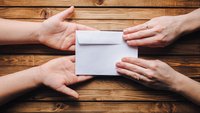 Ein Paar Hände liegt mit den Handflächen nach oben auf einem Holztisch und nimmt vorsichtig einen Briefumschlag eines anderen Paars Hände entgegen.