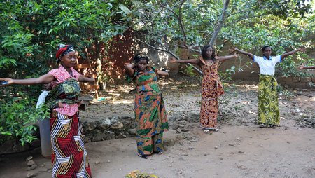 Four women participating in physical exercises outdoors, in a courtyard with trees and bushes providing shade. 